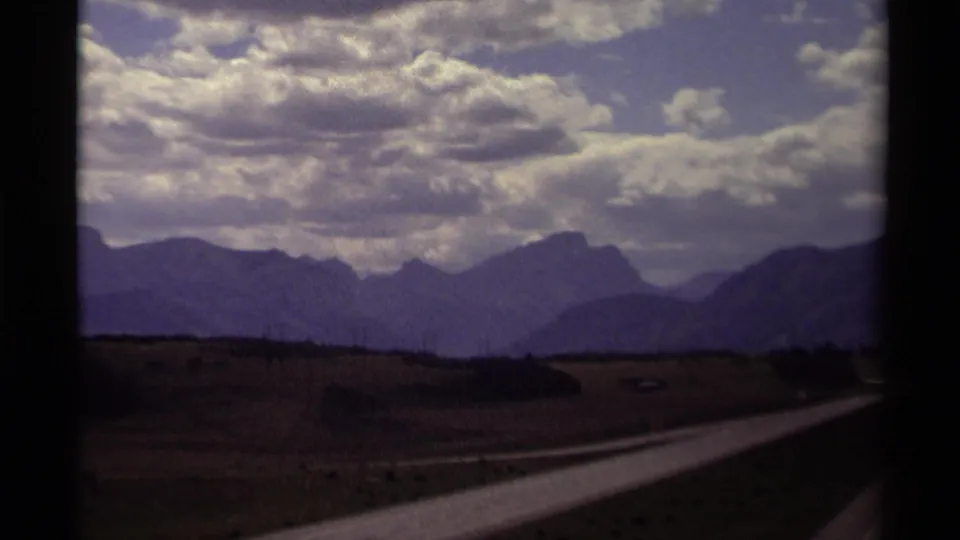 8mm home-movie frame of roadside fields, a highway edge, and distant Canadian mountains under thick cloud cover.