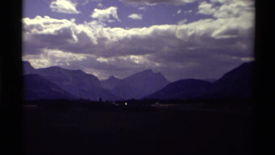 Canadian mountain landscape on 8mm film, with distant peaks, heavy clouds, and a dark foreground frame suggesting a moving travel viewpoint.