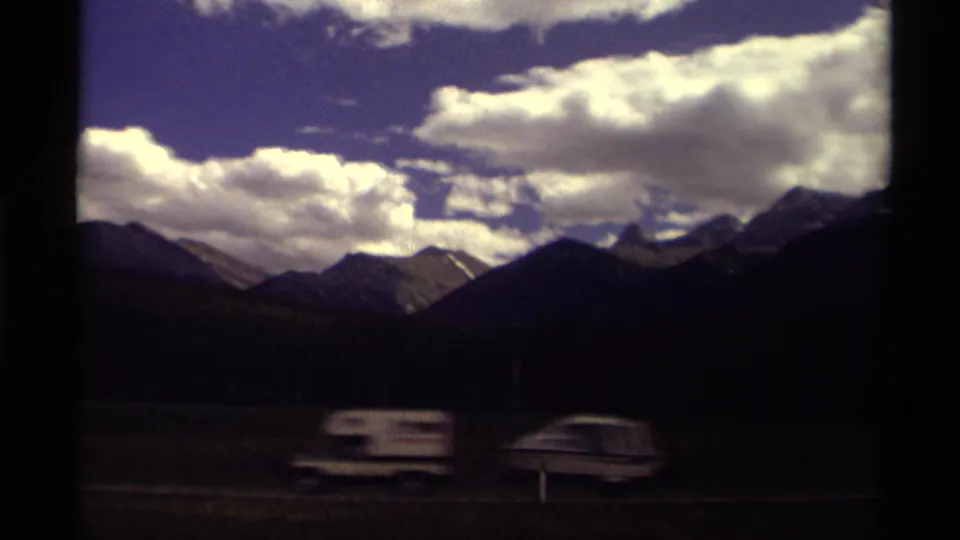 1979 Canadian road-trip frame with motion-blurred vehicles in the foreground and mountain silhouettes under a bright clouded sky.