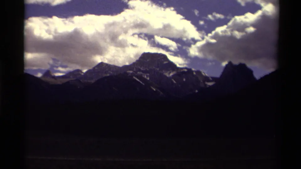 1979 Canadian home-movie frame showing rugged mountains beneath bright clouds, bordered by dark film or window edges.