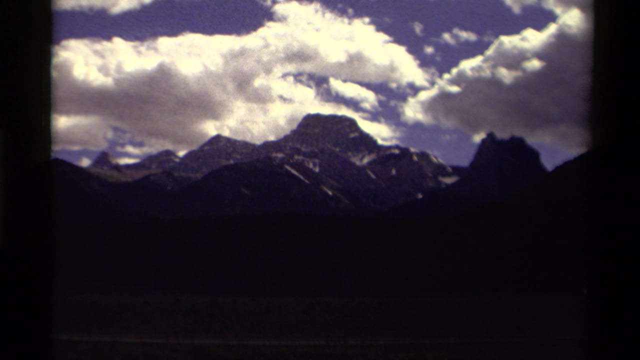1979 Canada 8mm frame showing dark mountain silhouettes and snow-streaked peaks under a bright, clouded sky.