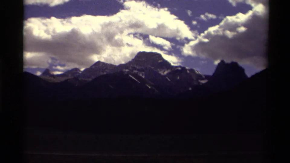 1979 Canada 8mm frame showing dark mountain silhouettes and snow-streaked peaks under a bright, clouded sky.