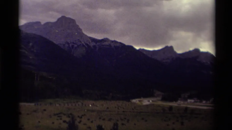 1979 Canada 8mm frame looking across a mountain valley toward a road, scattered buildings, and dark ridges below heavy clouds.