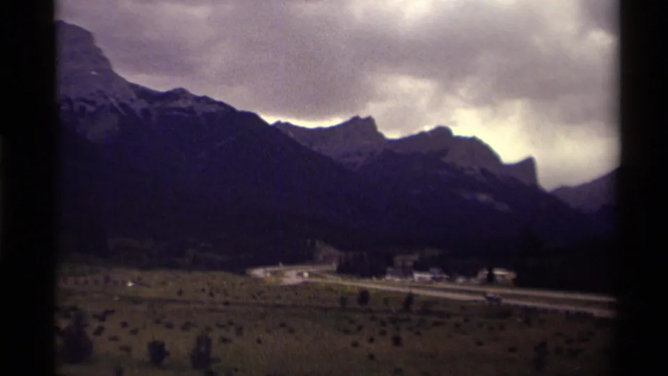 1979 Canada 8mm frame showing roadside buildings and an open valley foreground beneath dark mountain slopes and cloud cover.