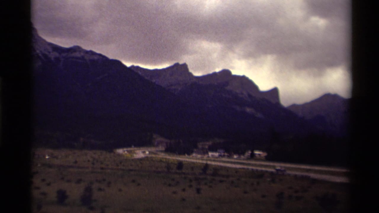 1979 Canadian 8mm frame showing a road through a mountain valley under heavy clouds, seen through a dark window or gate edge.