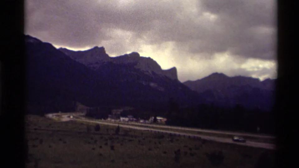 Canadian mountain road view from 1979 with small cars traveling along a highway, valley buildings, and heavy cloud cover.
