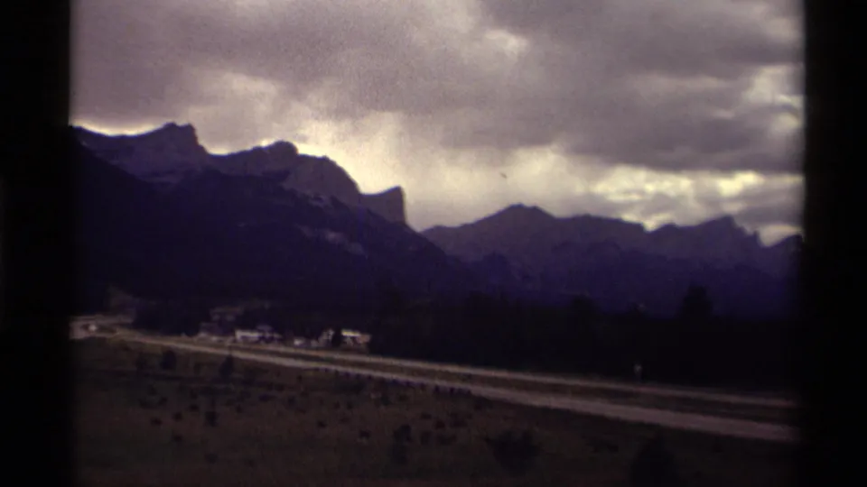8mm travel frame of a Canadian mountain valley in 1979, showing a road, low buildings, and shadowed peaks under dramatic clouds.