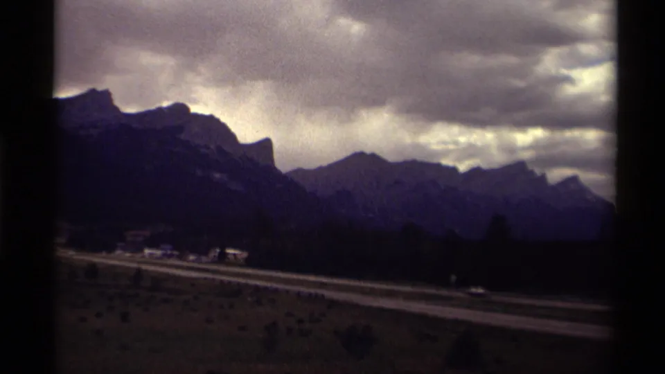Final 1979 Canadian 8mm frame with a highway, small cars, dark tree line, and mountains under a textured overcast sky.