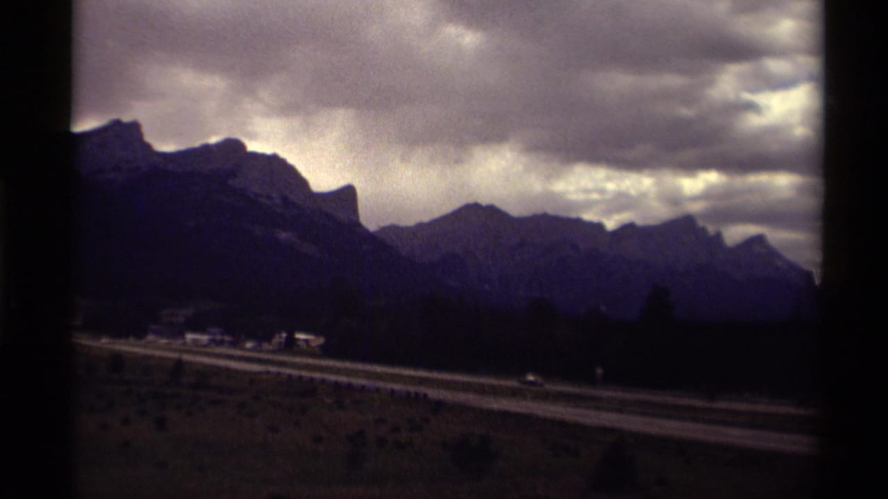 1979 Canadian home-movie frame showing a mountain range, dramatic clouds, and a highway in the valley below.