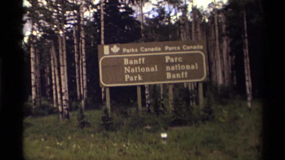Close visitor-shot view of the bilingual Banff National Park sign in Canada, surrounded by trees and low vegetation.
