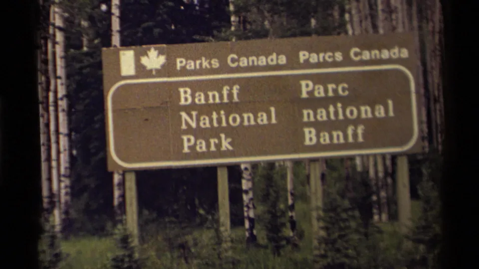 1979 8mm frame showing the Banff National Park sign large in view, with bilingual text and dense birch forest behind.