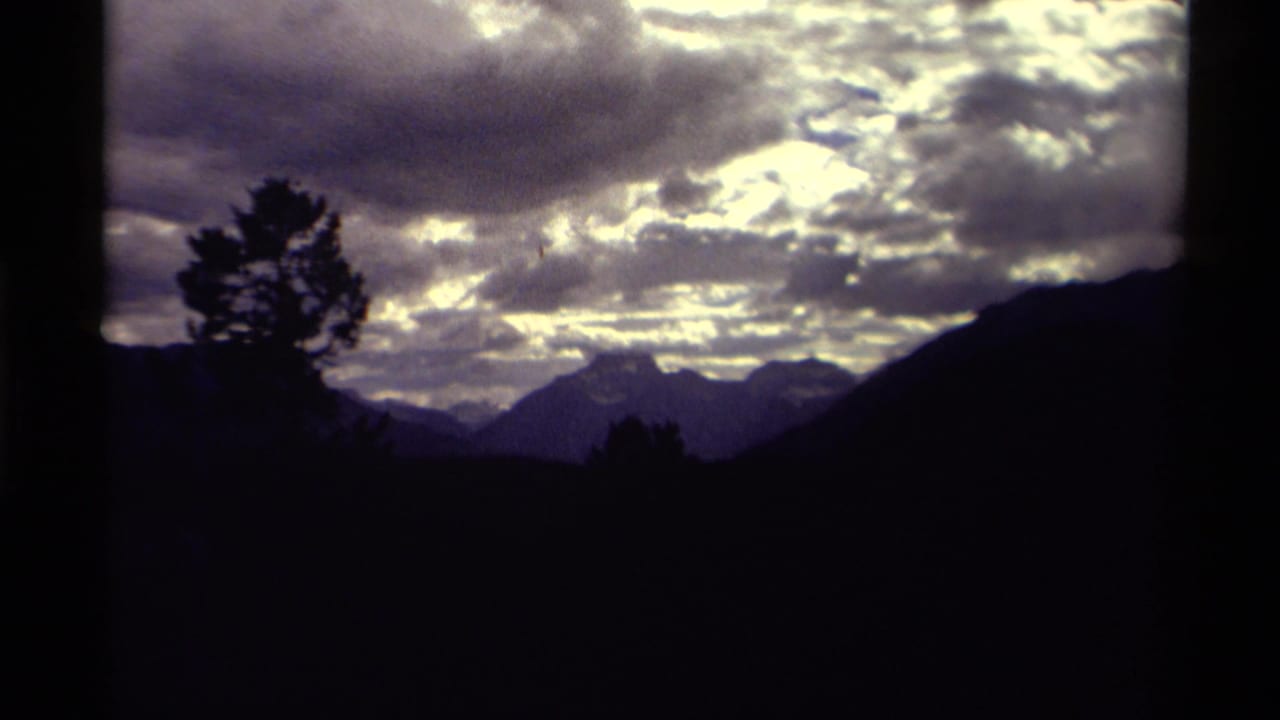 1979 Canada 8mm frame showing silhouetted evergreens and distant mountains under layered, luminous clouds.