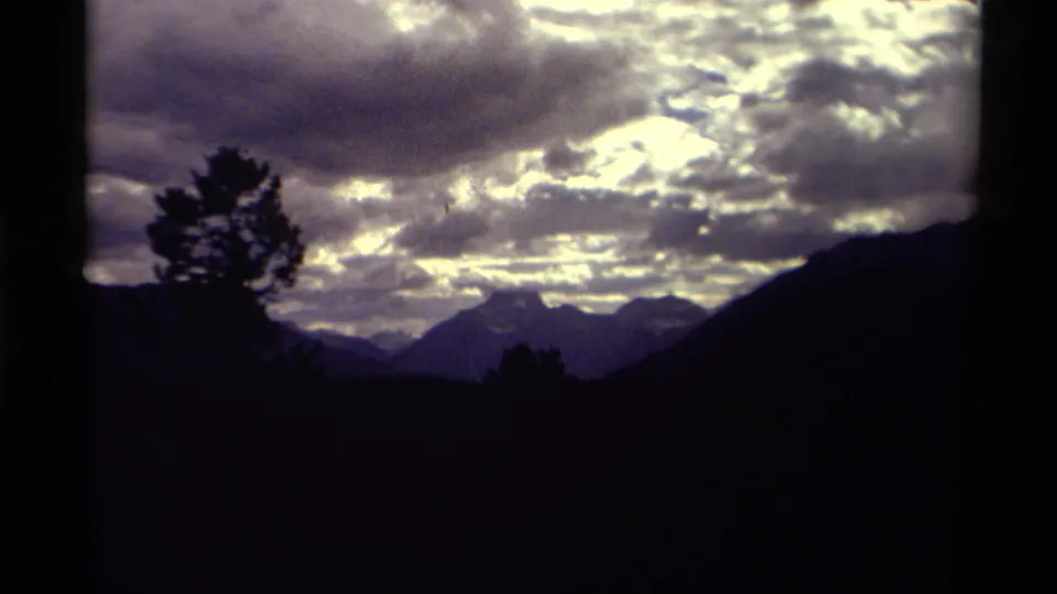 1979 Canada 8mm frame showing silhouetted evergreens and distant mountains under layered, luminous clouds.