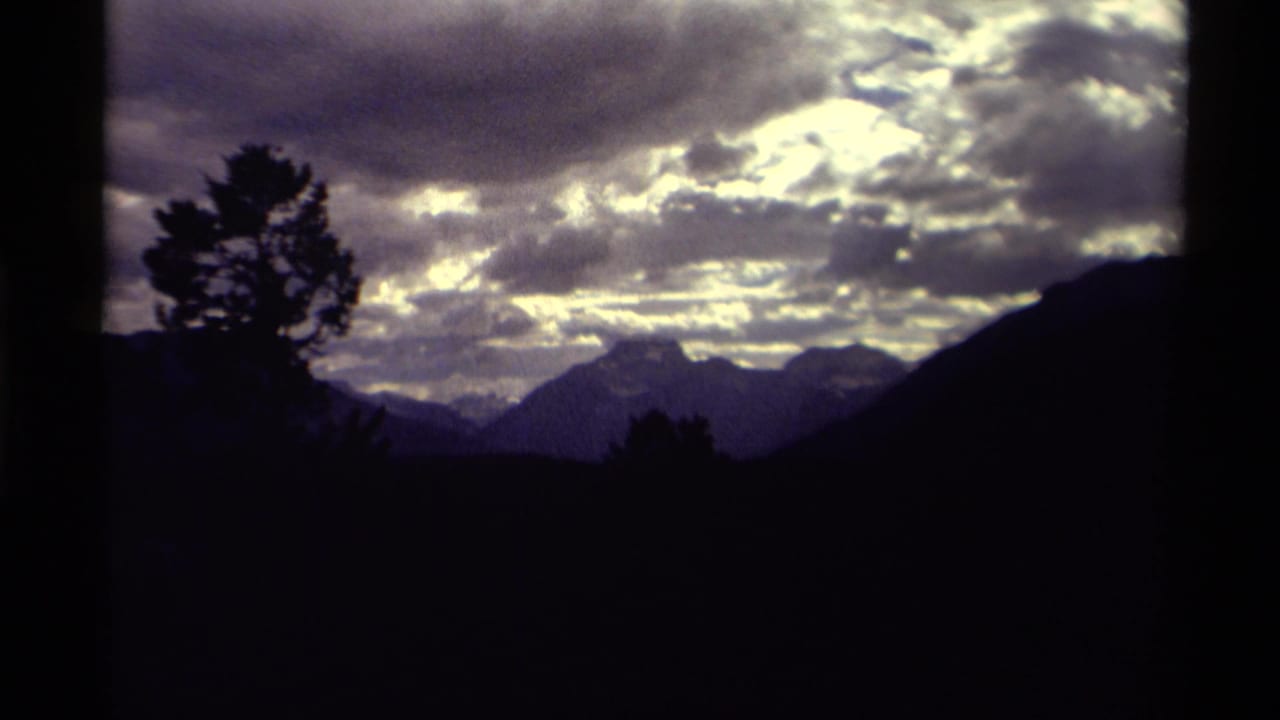 Vintage 8mm view of a Canadian mountain horizon, framed by a dark tree and glowing cloud cover.
