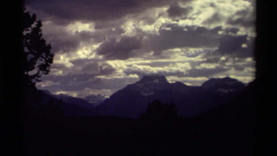1979 Canadian mountain scenery on 8mm film, with dark peaks, layered cloud cover, and tree silhouettes at the frame edges.