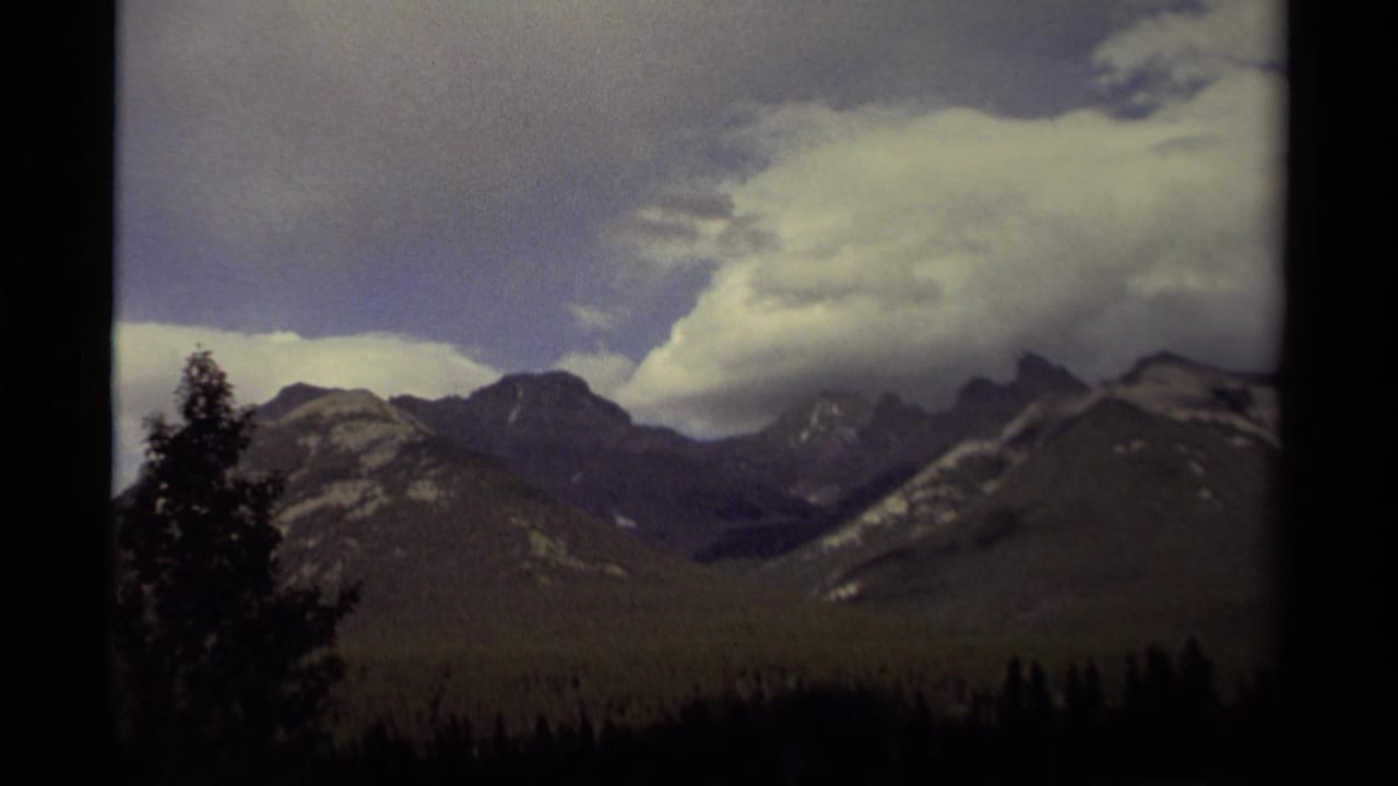Wide mountain landscape with dark trees, forested slopes, snow-dusted peaks, and cloudy sky.