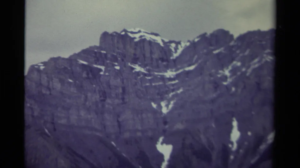Zoomed-in 1979 Canada 8mm frame showing rugged gray-blue mountain faces with lingering snow patches below a muted sky.