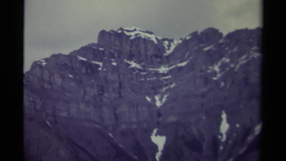 Close 8mm view of a Canadian mountain ridge, showing jagged rock texture, streaks of snow, and soft archival color under cloud cover.
