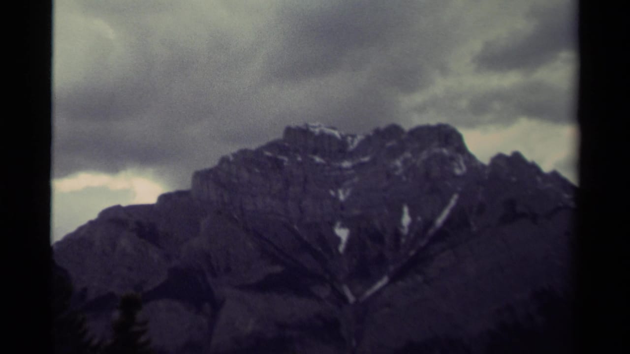 Rocky mountain ridge with snow streaks and heavy clouds, framed by dark film edges.