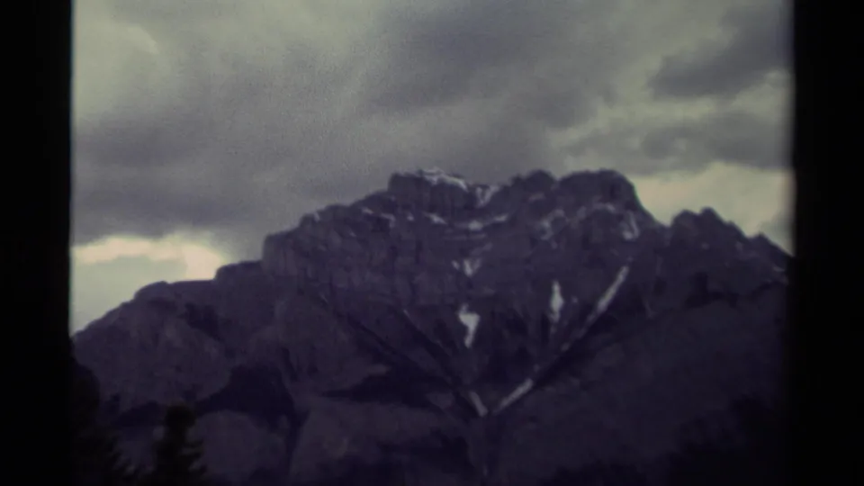 The zoom begins to reveal more of the Canadian mountain ridge, with snow markings and an overcast sky in soft 8mm color.