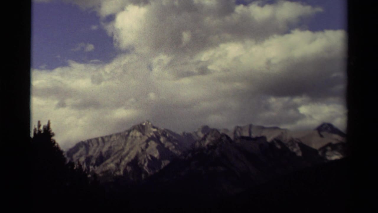 1979 Canadian 8mm view of jagged, snow-dusted mountains beneath a partly cloudy blue sky, framed by dark trees.