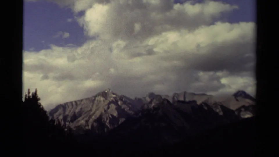 1979 Canadian 8mm view of jagged, snow-dusted mountains beneath a partly cloudy blue sky, framed by dark trees.