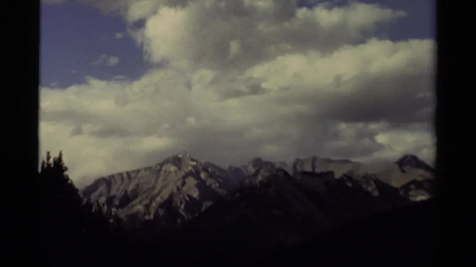 Canadian 8mm mountain landscape from 1979, showing distant rocky peaks, heavy clouds, blue sky, and silhouetted evergreens.