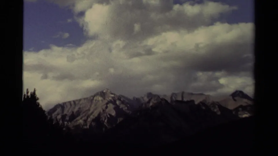 Late-1970s 8mm Canada travel footage shows clouds gathering above snow-marked mountains and dark roadside evergreens.