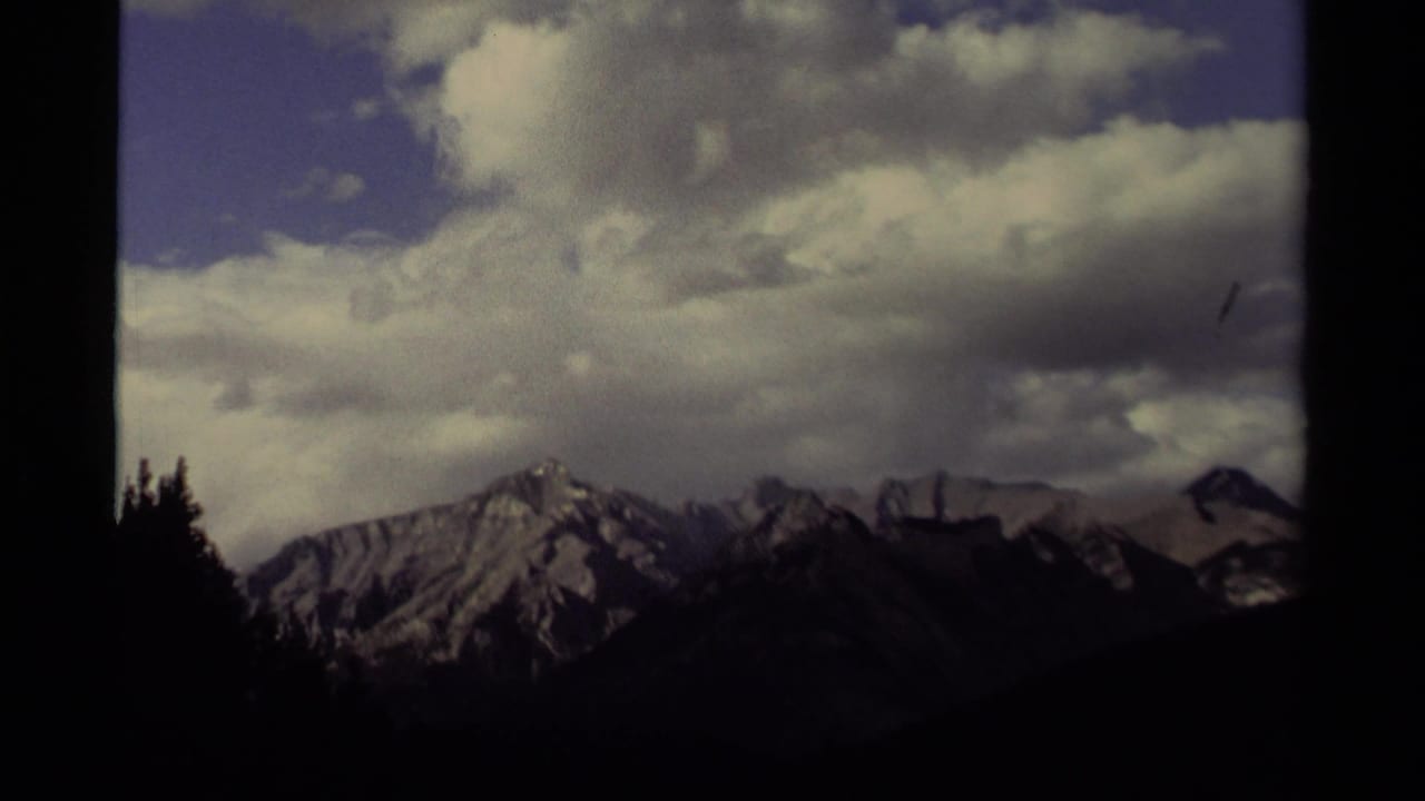 1979 Canadian 8mm frame showing a distant snowy mountain ridge, dark pine forest edge, and a large cloudy sky.