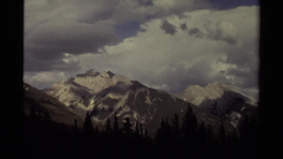 1979 Canadian 8mm frame of snow-capped mountain peaks above a dark pine forest with bright and shadowed clouds overhead.