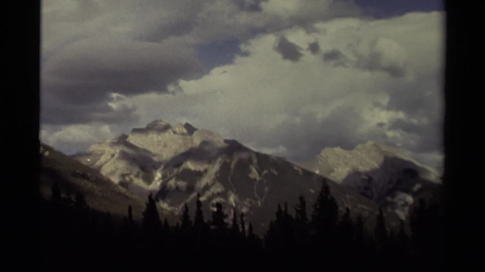 1979 Canadian 8mm frame with a snow-marked mountain face, distant ridge, dark conifers, and broad cloud cover.