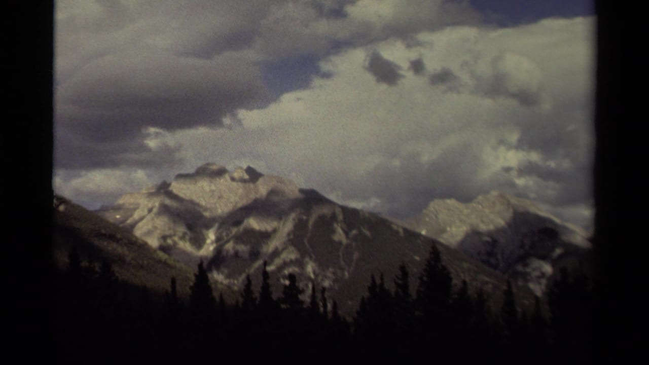 1979 Canada 8mm frame showing distant gray mountains with snow patches, heavy clouds, and a silhouetted evergreen forest foreground.