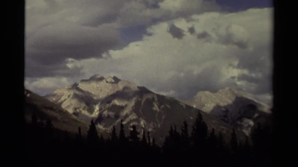 1979 Canada 8mm frame showing distant gray mountains with snow patches, heavy clouds, and a silhouetted evergreen forest foreground.