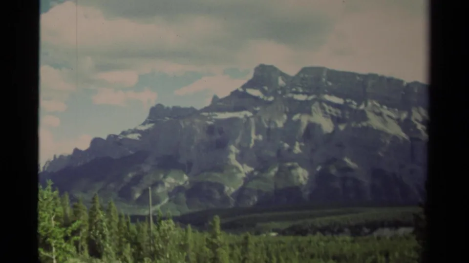 1979 Canada 8mm frame of a broad gray mountain face with white snow streaks, green forest below, and pale clouds overhead.