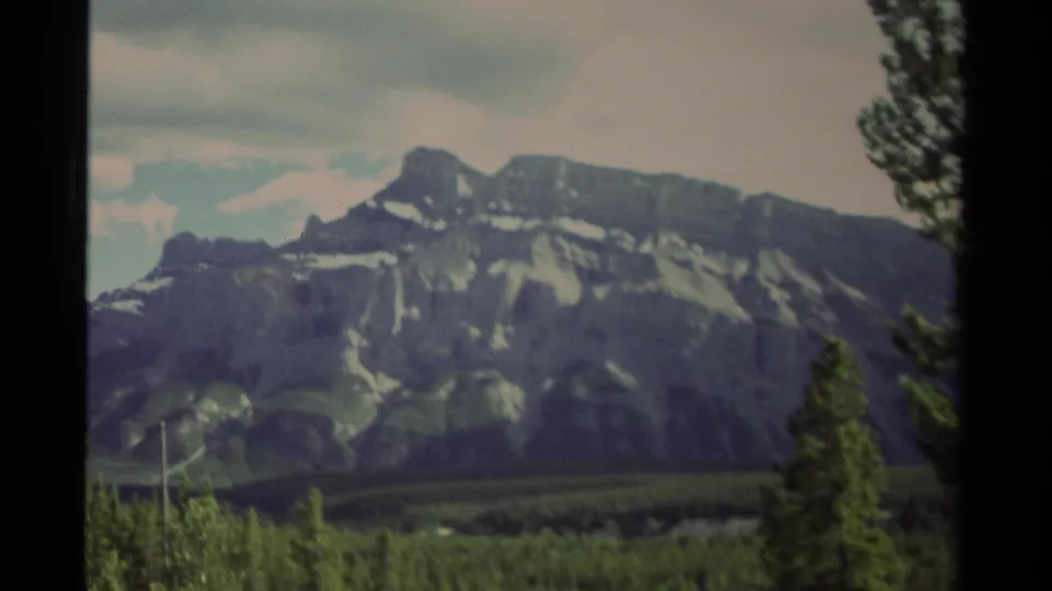 1979 Canada 8mm frame of a gray mountain with snow streaks, green forest foreground, cloudy sky, and a tree edge at right.