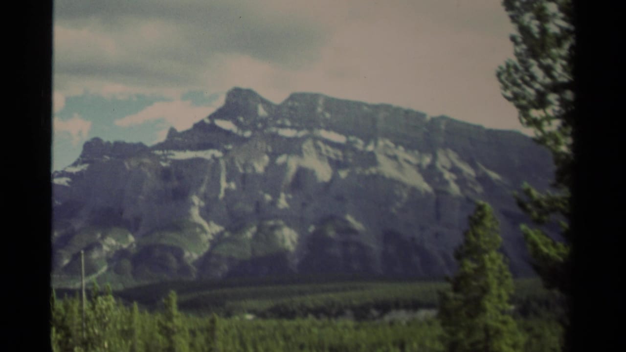 1979 Canadian 8mm frame showing distant rugged mountains, conifer forest, cloudy sky, and dark frame edges.