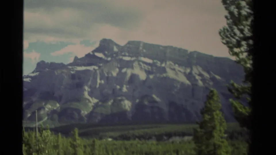 1979 Canadian 8mm frame showing distant rugged mountains, conifer forest, cloudy sky, and dark frame edges.