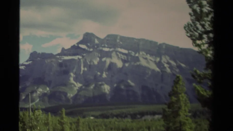Canadian wilderness view on 8mm film, with a rugged mountain ridge, conifers, cloudy sky, and vertical frame border.