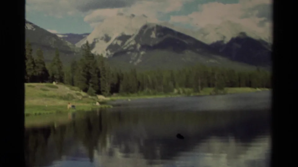 1979 Canadian 8mm lake scene with mountain backdrop, evergreen shoreline, grassy bank, and water reflections.
