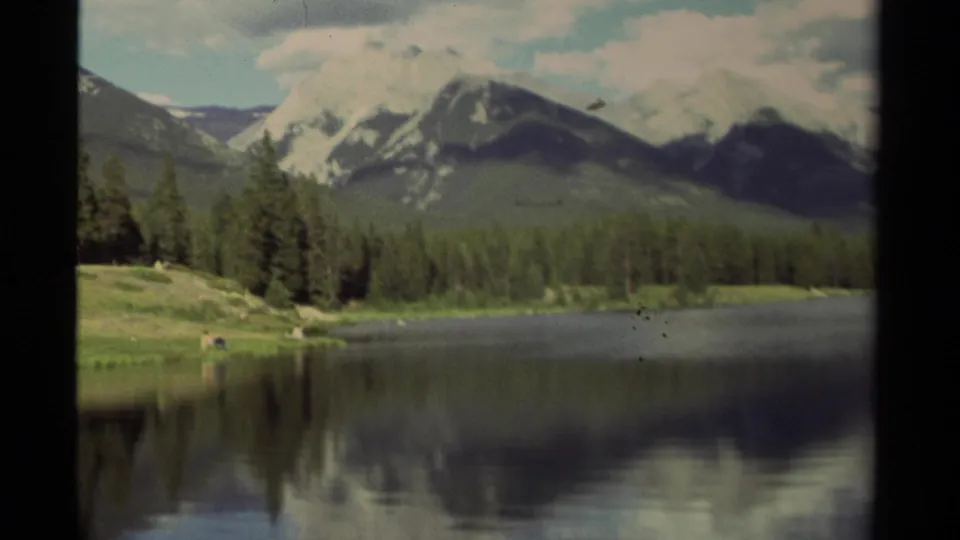 Canadian 8mm travel frame showing a reflective lake, conifer forest, and snow-streaked mountains beneath broken cloud.