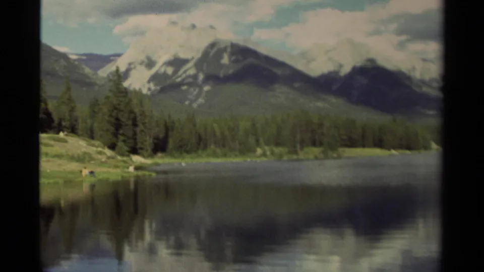 1979 Canadian 8mm frame showing snow-capped mountains behind evergreens, mirrored on a still lake under clouds.