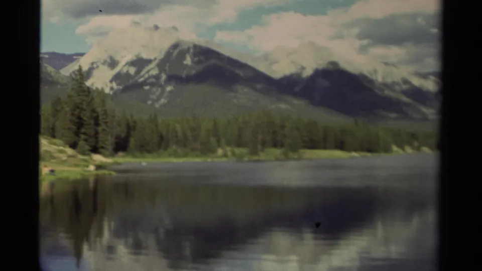 1979 Canadian 8mm view of an evergreen-lined lake with snow-streaked mountains and soft cloud shadows.