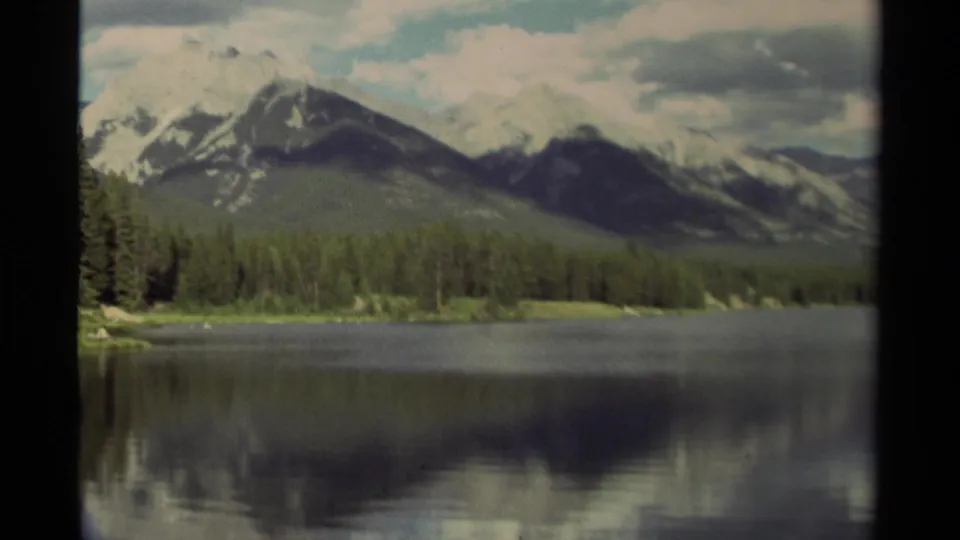 1979 Canadian landscape frame with a forested lakeshore, snow-marked peaks, and dark reflected shapes on the water.