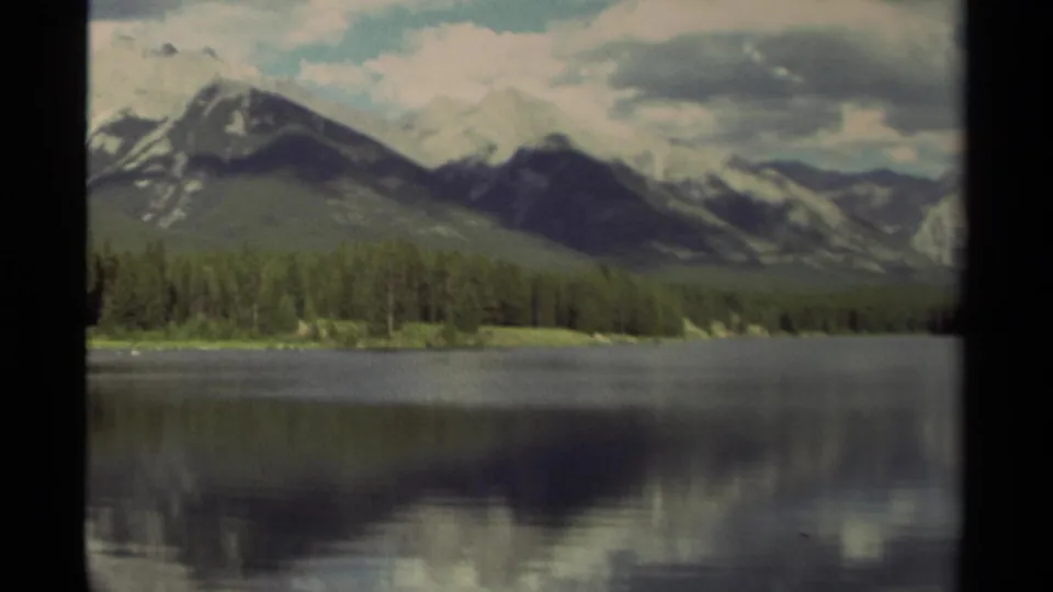 1979 Canadian 8mm frame holding on mountains, evergreen trees, and a broad lake reflection beneath a cloudy sky.