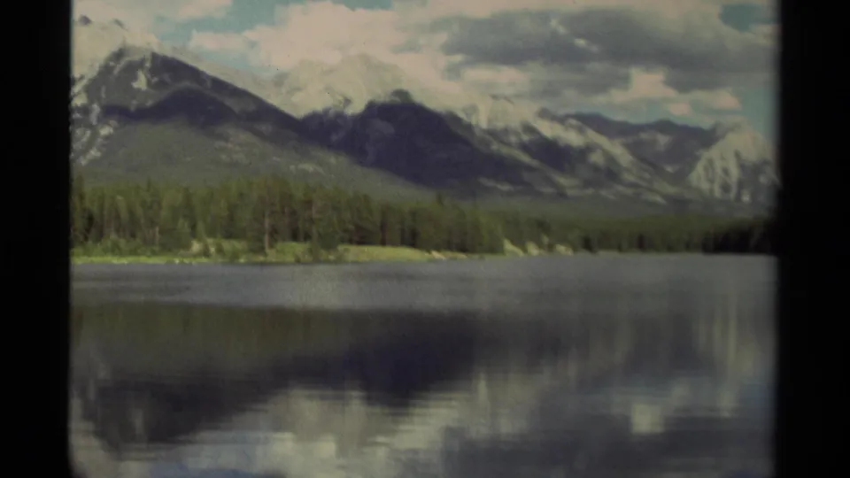 1979 Canada 8mm frame showing a still lake, evergreen shoreline, snow-dusted mountains, and soft cloud reflections.