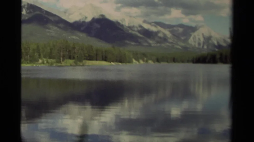 1979 Canada 8mm frame with a broad lake surface reflecting sky beneath a forested shoreline and mountain range.