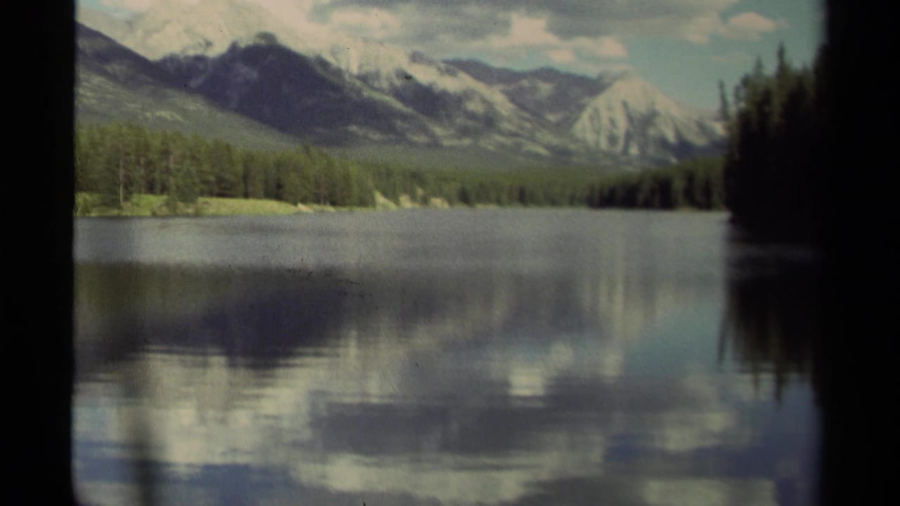 Calm lake reflecting forested mountains and cloudy sky.