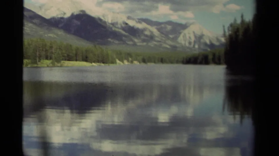 1979 Canadian 8mm frame of a calm mountain lake with evergreen shoreline, distant peaks, and cloudy reflections on the water.