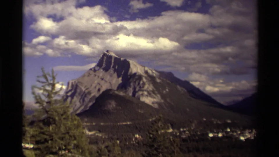 1979 Canadian 8mm frame with a prominent rocky mountain, foreground trees, a distant town, and bright clouds crossing the sky.