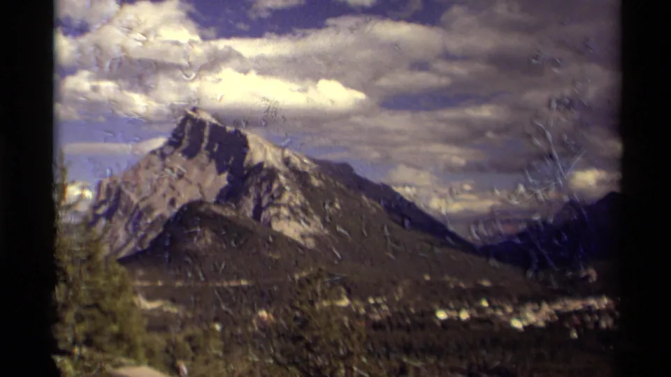 1979 Canadian 8mm frame of a mountain over a valley, with evergreen foreground, cloudy sky, and visible marks from the film scan.
